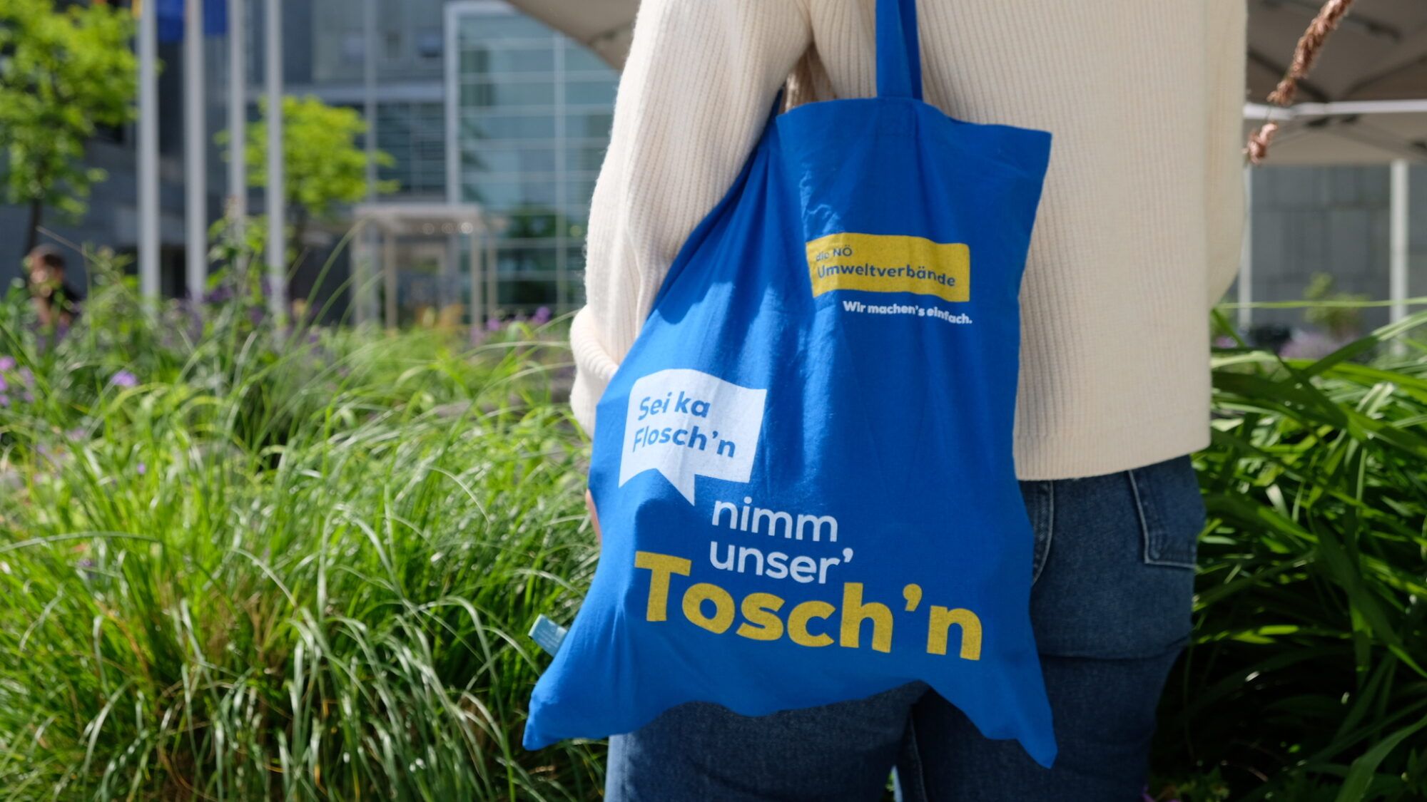 Eine Person tr&auml;gt eine blaue Stofftasche der N&Ouml; Umweltverb&auml;nde mit dem Aufdruck &bdquo;Sei ka Flosch&rsquo;n, nimm unser&rsquo; Tosch&rsquo;n&ldquo; in einer begr&uuml;nten, modernen Au&szlig;enanlage.