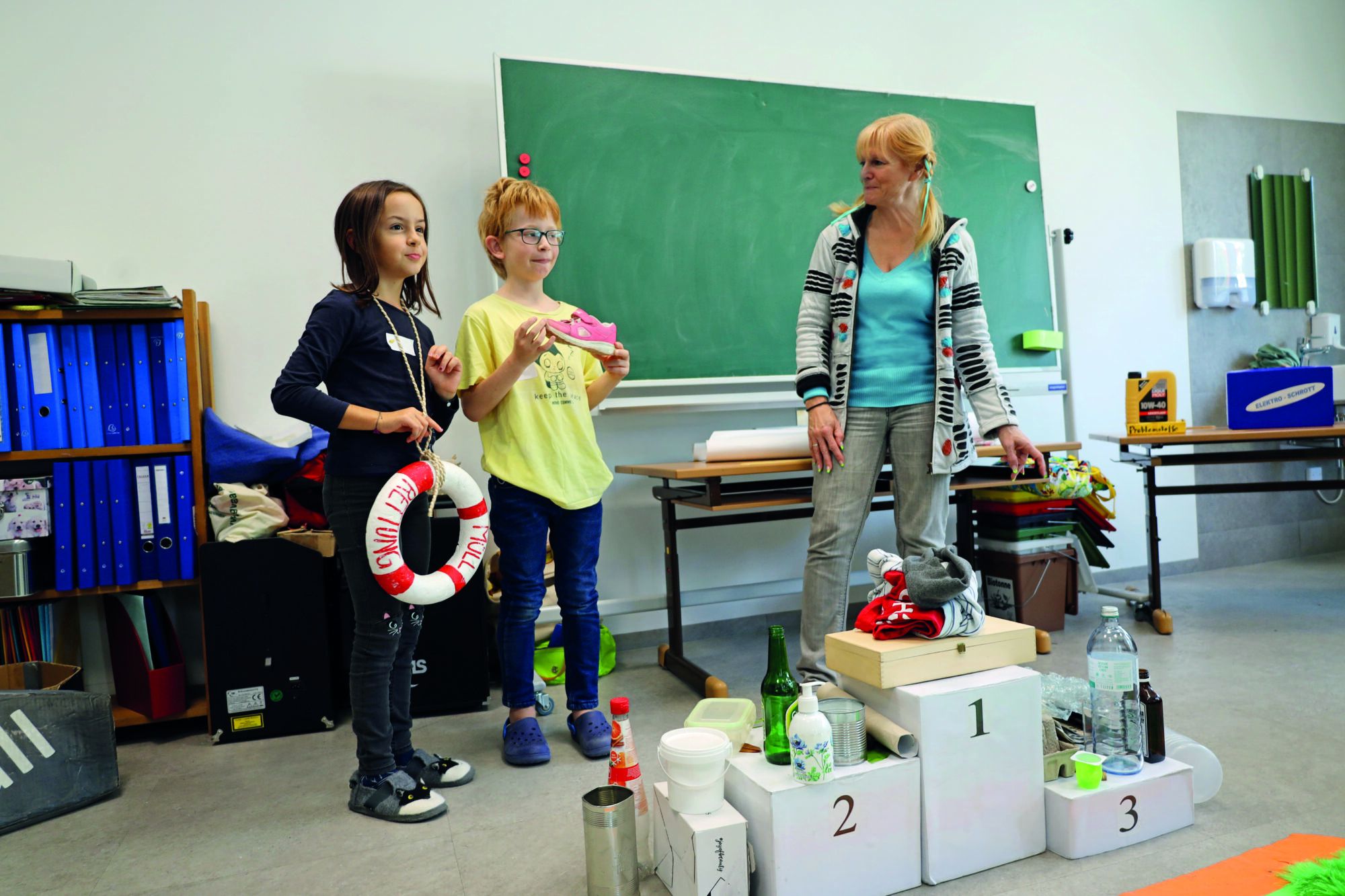 Zwei Kinder im Volksschulalter stehen neben einer bloden Frau mit geflochtenen Zöpfen und einer Schleife im Haar. Davor ein Siegespodest mit Abfällen darauf und im Hintergrund eine Tafel und ein Regal mit blauen Ordnern. 