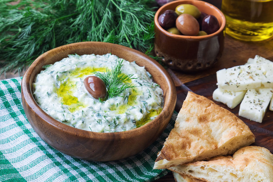 Cremiger Tsatsiki in einer rusitkalen braunen Keramischüssel, garniert mit Olivenöl und einer Olive. Daneben Weißbrot, Feta und Oliven.